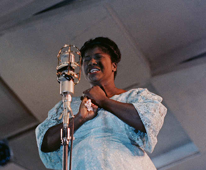 A woman shown waist up, holding her hands with her eyes closed, singing into a microphone.