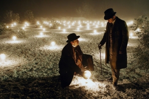 two men standing in a field with glowing bulbs, one man is holding a bulb looking up at the other man